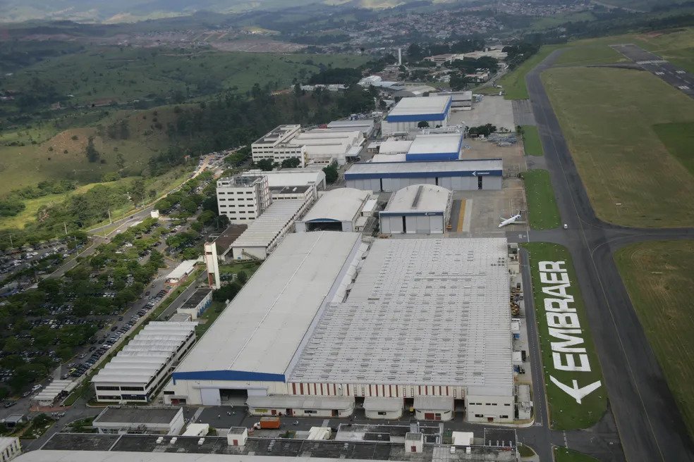 Vista aérea de complexo industrial da Embraer com hangares e pista de aeroporto, representando infraestrutura tecnológica e oportunidades para investir no Brasil. Vista aérea de complexo industrial da Embraer com hangares e pista de aeroporto, representando infraestrutura tecnológica e oportunidades para investir no Brasil.